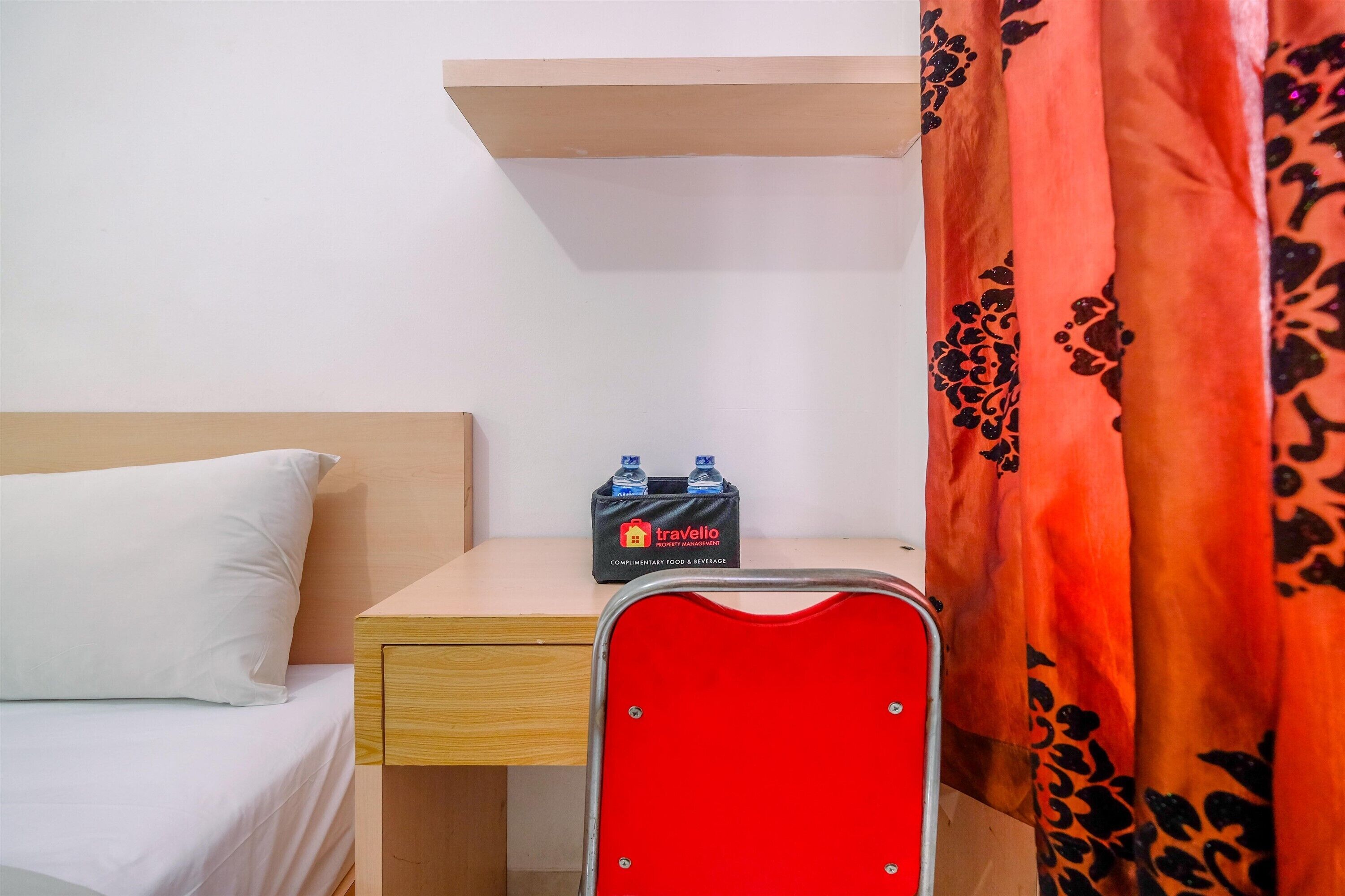 A hotel room with a bed, a wooden nightstand, a red chair, a small table with a minibar, and a red curtain with a floral pattern.