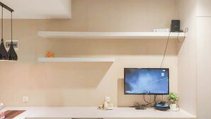 A modern office space with a white desk, a computer monitor, and a shelf with a speaker and a small plant. - Good and Homey Studio at Student Park Yogyakarta Apartment (Depok)