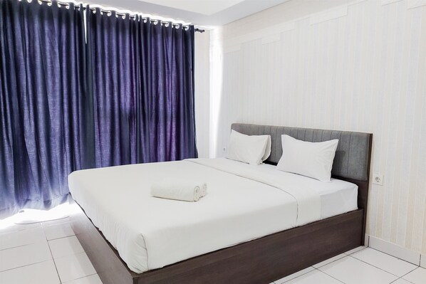 A neatly made bed with white linens and a rolled towel, a dark wooden headboard, and a window with dark curtains. - Cozy and Enjoy Living Studio Casa De Parco Apartment (Pagedangan)