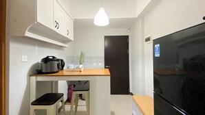 A compact kitchen with a black refrigerator, a countertop with a rice cooker, a small stool, and a potted plant. - Minimalist And Cozy 1BR at Royal Heights Apartment (Bogor)
