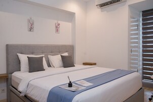 A neatly made bed with a grey headboard, white and grey pillows, and a blue bedspread. There are two wall-mounted artworks above the bed, and a small wooden nightstand beside it.