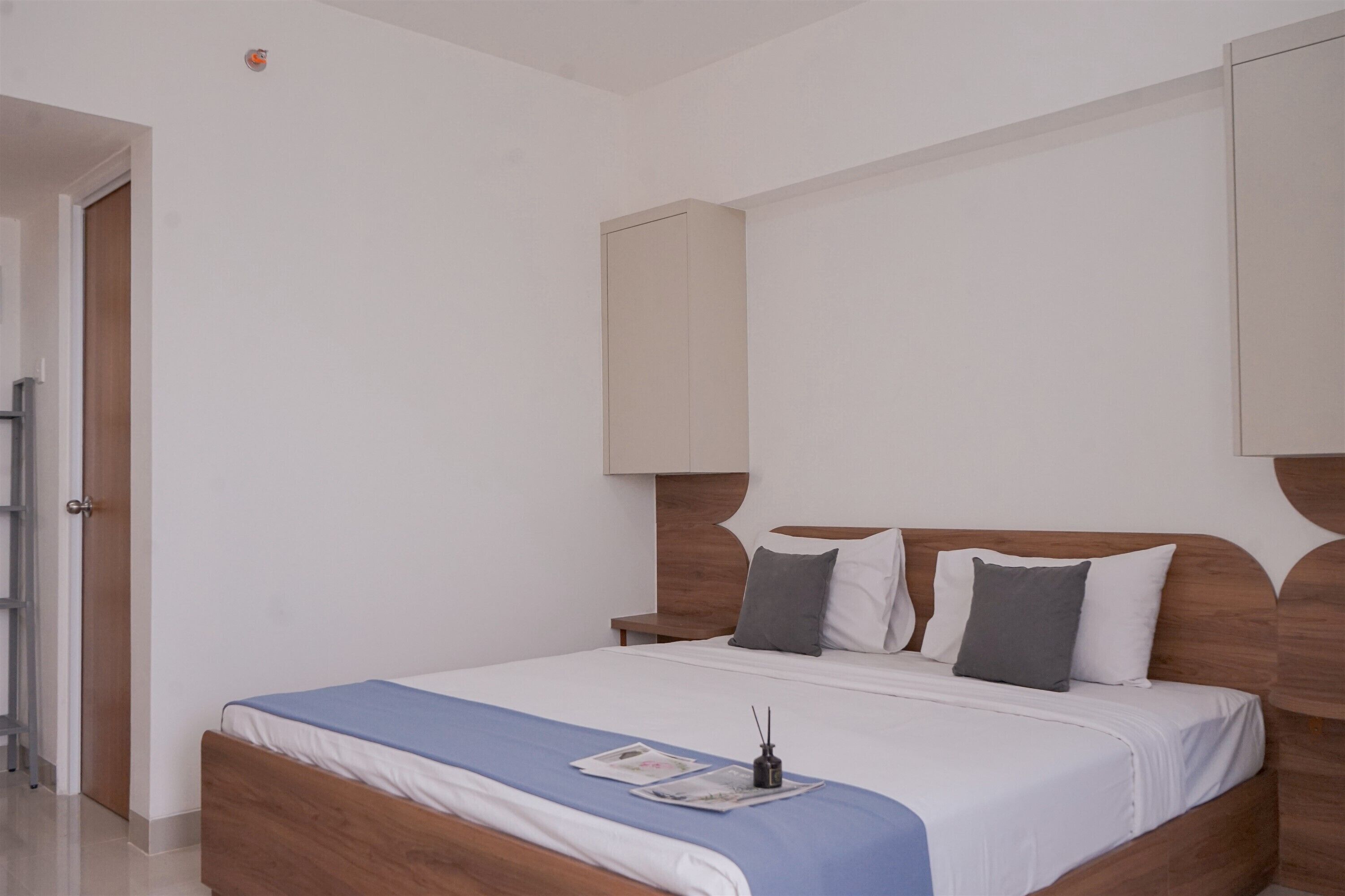 A modern bedroom with a wooden bed, white bedding, and a blue throw blanket. There is a nightstand with a lamp and a small table beside the bed.