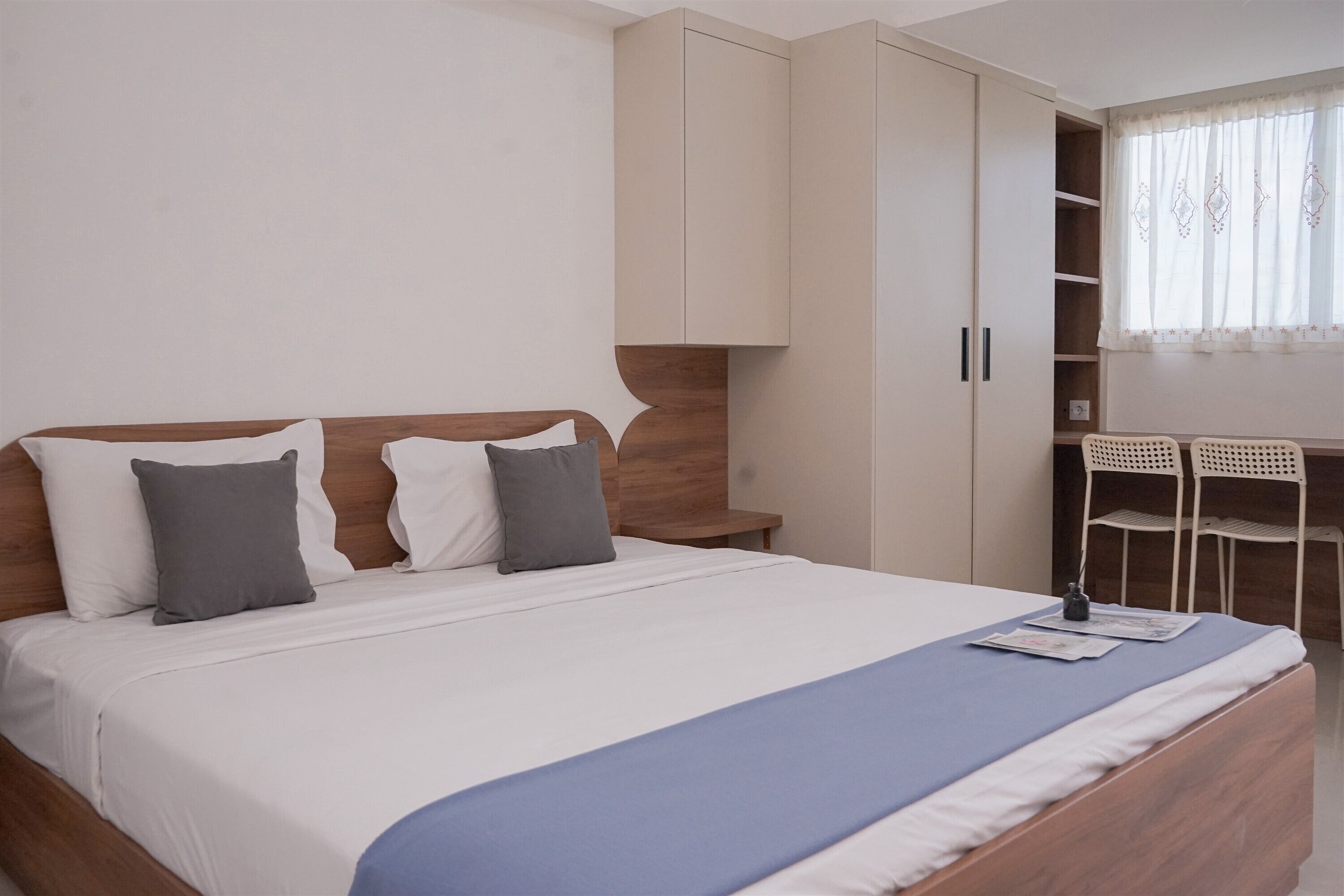 A bedroom with a large bed, a wooden headboard, a wardrobe, a desk with chairs, and a window with curtains.