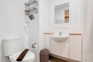A modern bathroom with a toilet, sink, mirror, and a trash bin. - Comfy 1BR Apartment at Tamansari Skylounge Makassar (Makassar)