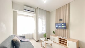 A modern living room with a grey sofa, a small white table, a TV mounted on a wooden panel wall, and an air conditioning unit on the ceiling. - Fully Furnished and Comfortable 2BR Nifarro Park Apartment (Jakarta)