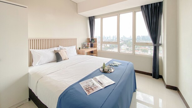 A hotel room with a large bed, a nightstand, a window with curtains, and a city view. - Fully Furnished and Comfortable 2BR Nifarro Park Apartment (Jakarta)