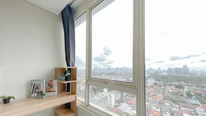 A room with a large window offering a city view, a wooden shelf with plants, and a framed picture. - Fully Furnished and Comfortable 2BR Nifarro Park Apartment (Jakarta)