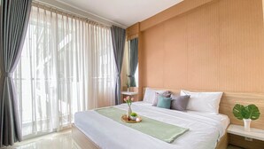 Een hotelkamer met een groot bed, een nachtkastje met een plant erop en uitzicht op de stad door dunne gordijnen.