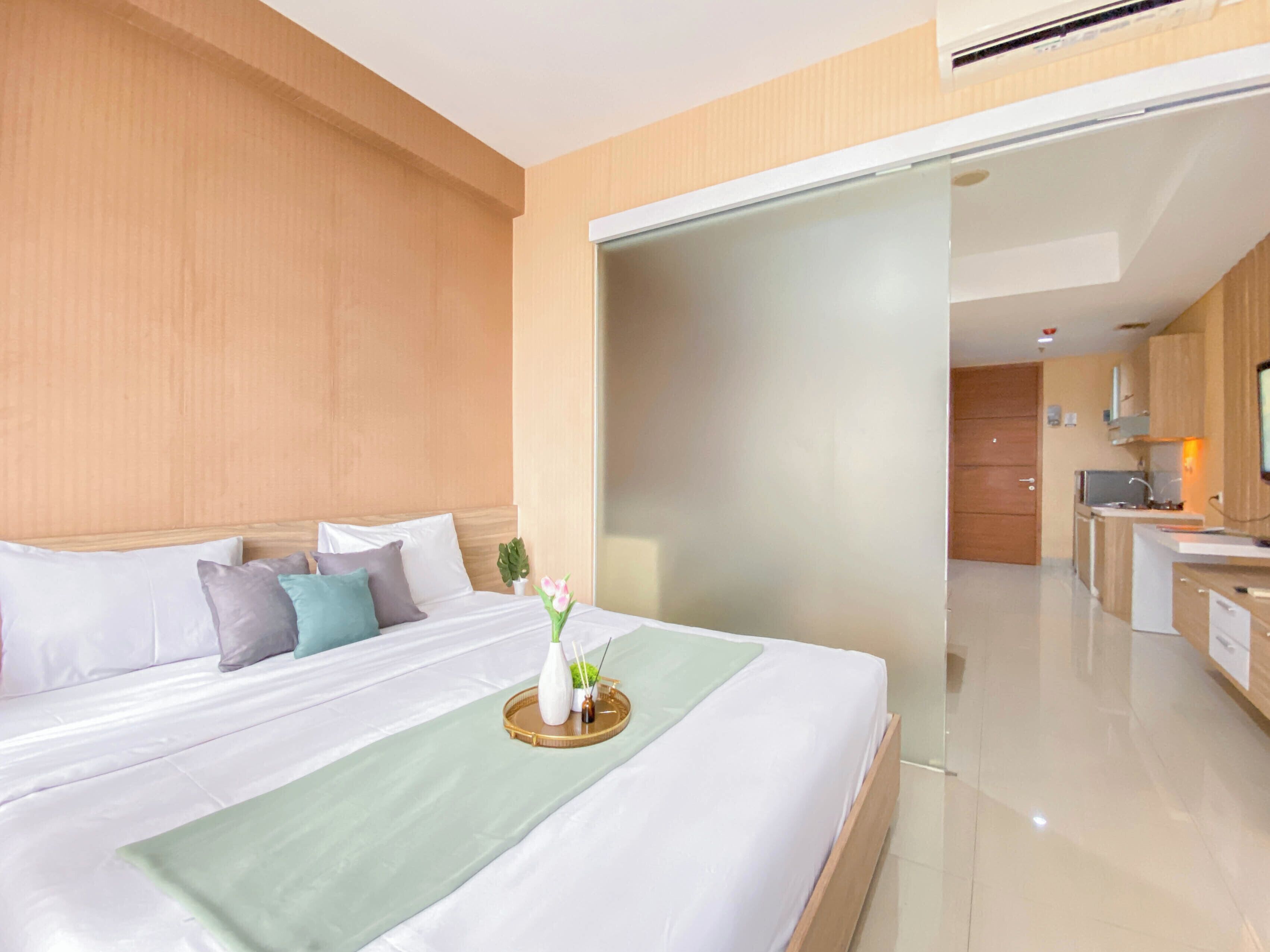 A modern hotel room with a large bed, a bedside table, a small potted plant, and a view into the kitchen area.