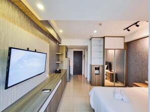A modern hotel room with a flat-screen TV mounted on the wall, a wooden desk, a bed with white linens, and a walk-in closet. - Cozy and Nice Studio at Student Castle Yogyakarta Apartment (Depok)