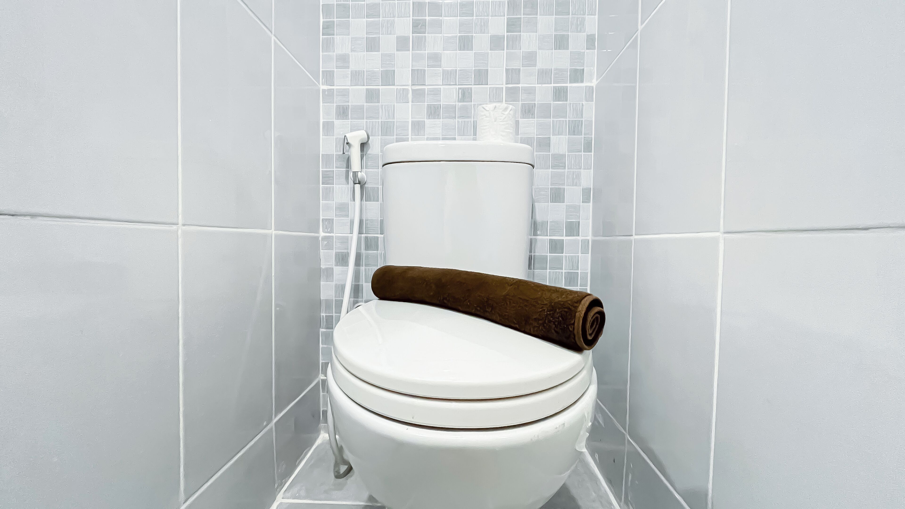 A white toilet with a brown towel on the seat, set against a tiled wall with a mosaic pattern.