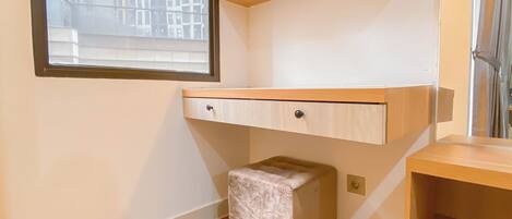 A corner room with a built-in desk, a small ottoman, and a window overlooking a building.