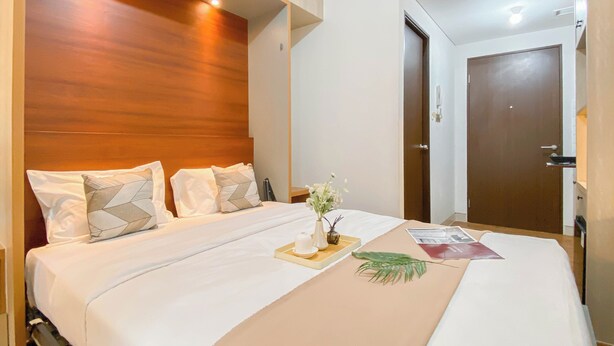 A neatly made bed with a wooden headboard, a bedside table with a vase of flowers, and a door to the right. - Good Place and Cozy Studio at Transpark Cibubur Apartment (Bekasi)
