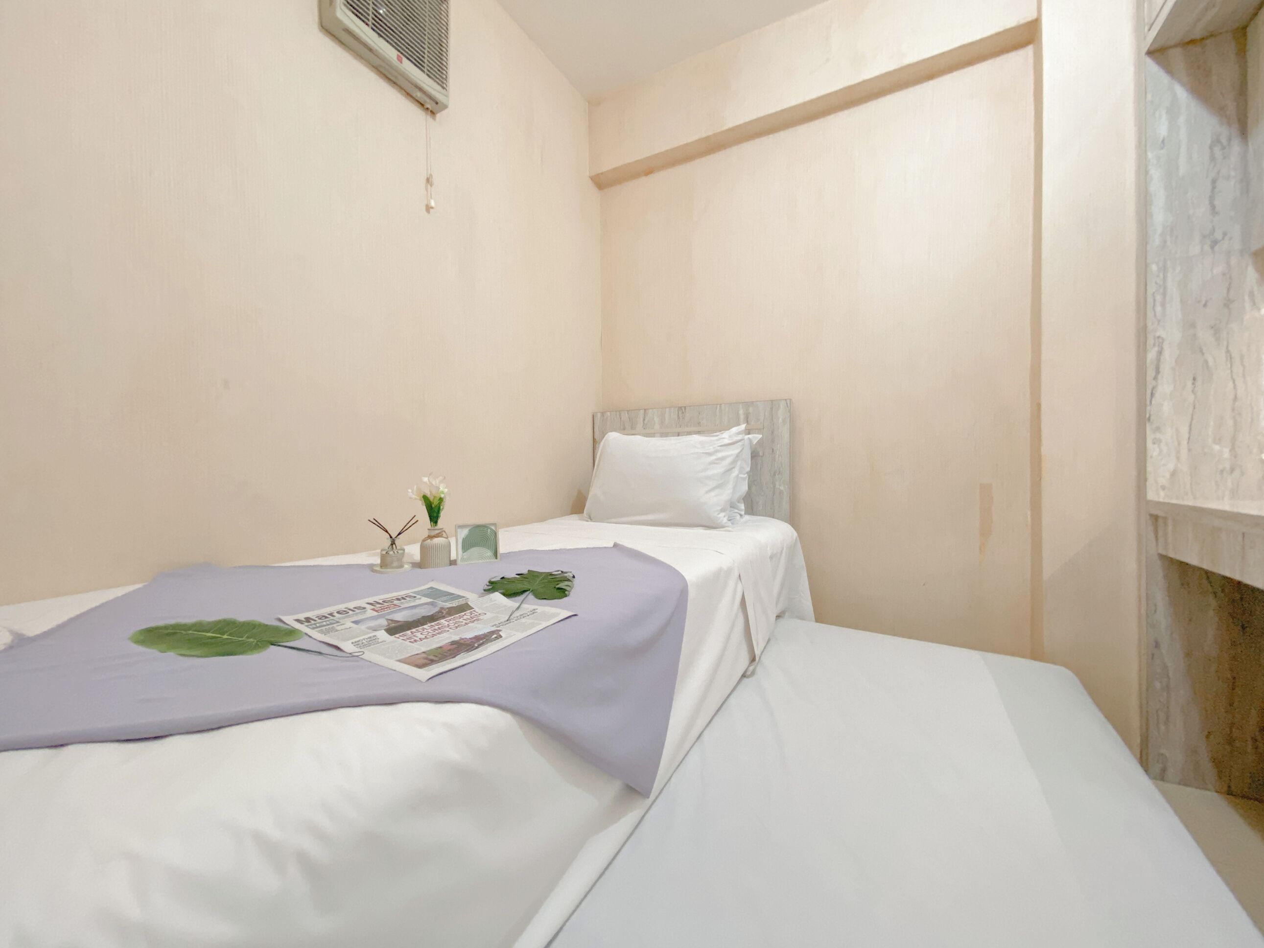 A single bed with a white headboard and a purple blanket. There is a small table with a plant, a newspaper, and a candle. The room has plain walls and a wooden shelf.