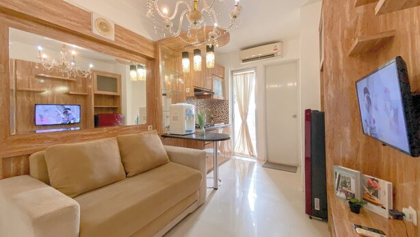 A modern living room with a beige sofa, a chandelier, a television, and a kitchen area with wooden cabinets and a coffee machine. - Best Deal and Modern Style 2BR Apartment Bassura City (Jakarta)