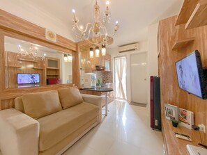 A modern living room with a beige sofa, a chandelier, a television, and a kitchen area with wooden cabinets and a coffee machine. - Best Deal and Modern Style 2BR Apartment Bassura City (Jakarta)