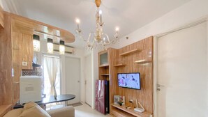 A living room with a sofa, a TV mounted on a wooden wall, a chandelier, and a kitchen area with cabinets and a refrigerator. - Best Deal and Modern Style 2BR Apartment Bassura City (Jakarta)