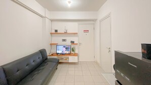 A modern living room with a black sofa, a television, a small table with a plant, and a white door with a fire safety sign. - Comfy 1BR Apartment with Extra Room at Green Pramuka City near Mall (Jakarta)
