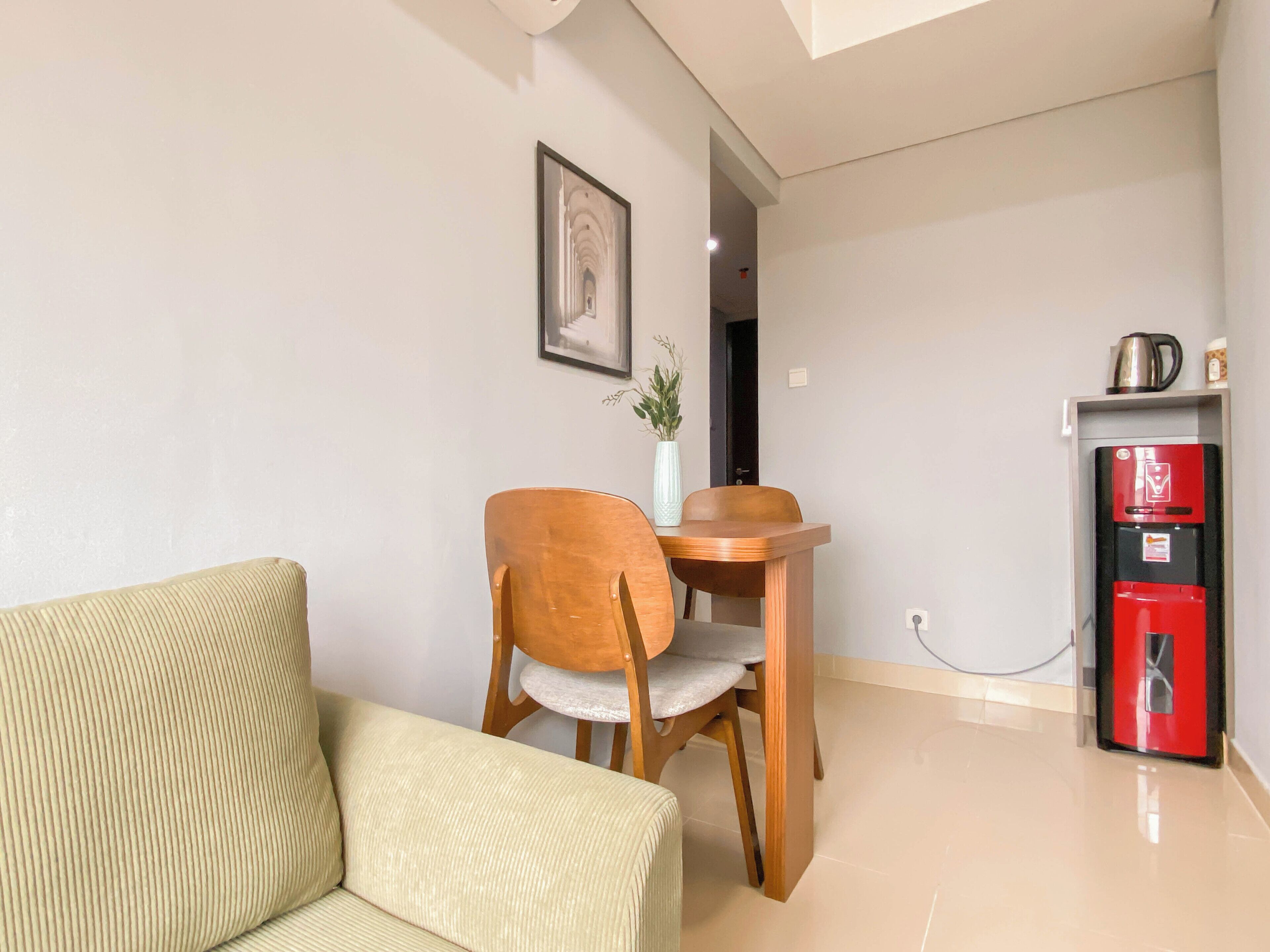 Un salon comprenant un canapé, une table à manger en bois avec deux chaises, un tableau encadré au mur, une petite plante et un appareil électroménager rouge.