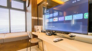 Ein moderner Arbeitsraum mit einem großen Monitor, der eine Google TV-Oberfläche anzeigt, einem bequemen Stuhl und einem Fenster mit Jalousien.