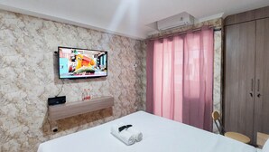 Une chambre d’hôtel avec un lit, un téléviseur fixé au mur, une étagère en bois, une armoire et un rideau rose.