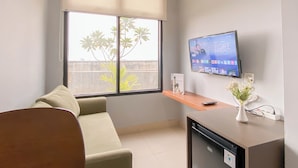 A modern living room with a sofa, a TV mounted on the wall, a small table with a vase, and a window with blinds. - Simply Look And Best Deal 2BR at Pollux Chadstone Apartment (Cikarang)