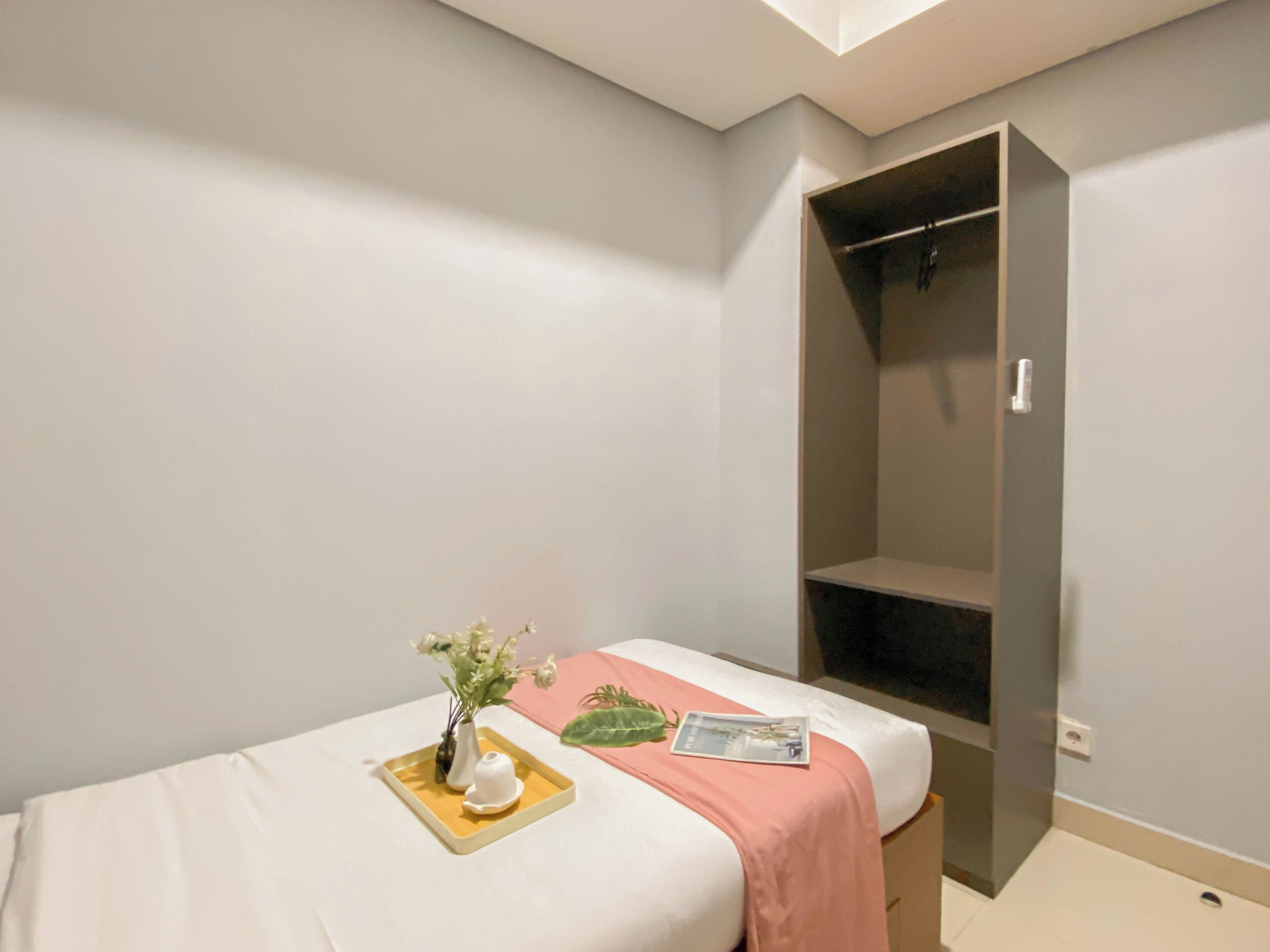 A bedroom with a single bed, a bedside table with a vase of flowers, a magazine, and a pink blanket. There is a wardrobe with a mirror and a door.