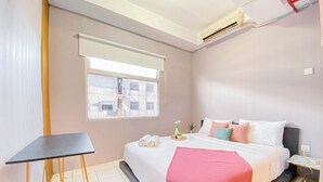 A neatly arranged hotel room with a bed, a small table, a window with a view of buildings, and an air conditioning unit on the wall. - Spacious 1BR at Grand Asia Afrika Apartment (Bandung)