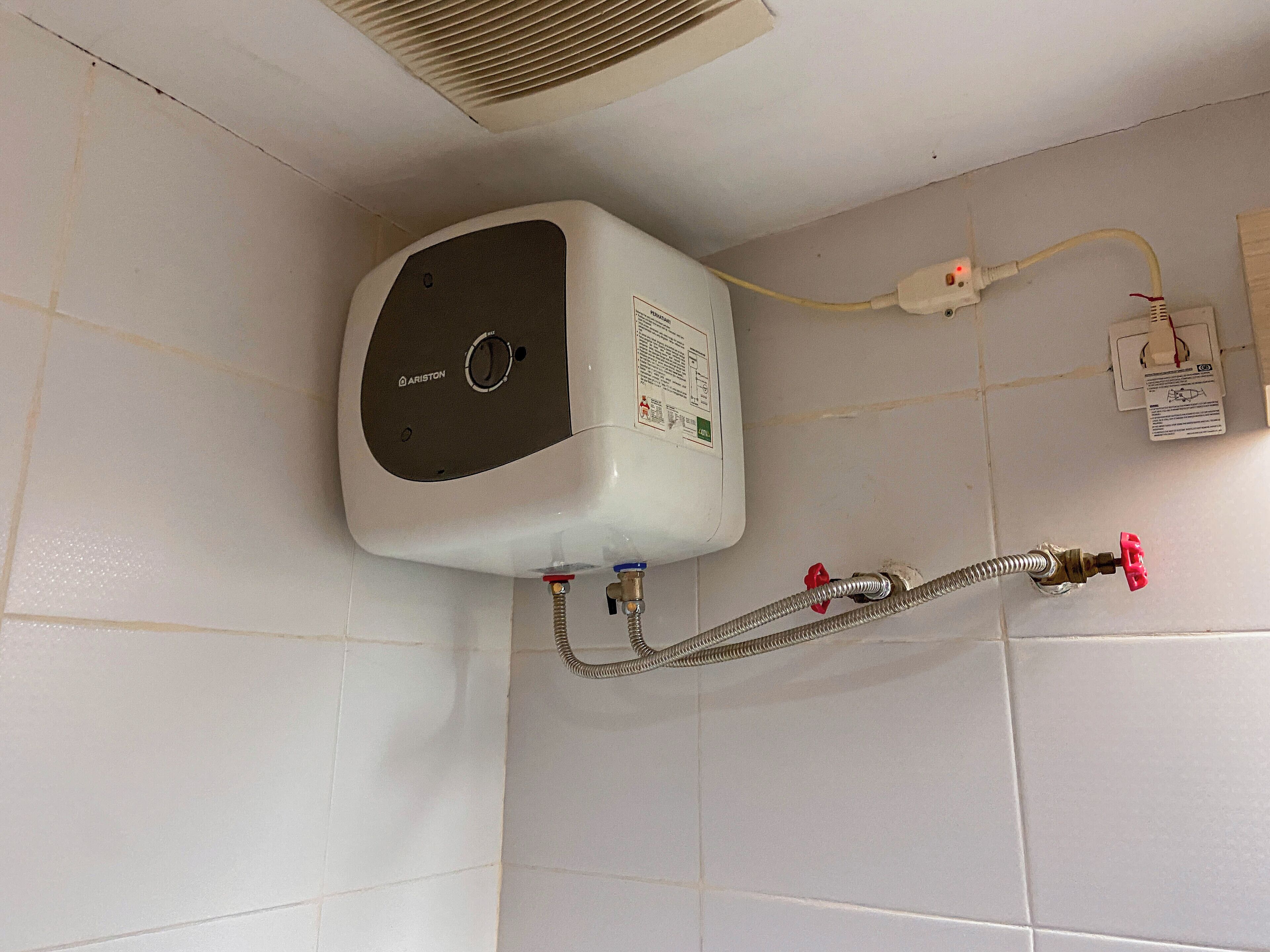 Un chauffe-eau blanc fixé sur un mur recouvert de carreaux, muni d’un bouton de commande, relié à un tuyau flexible et à un robinet rouge.