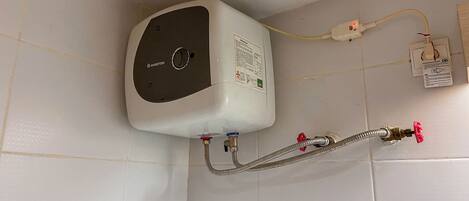 A white water heater mounted on a tiled wall with a control knob, connected to a flexible hose and a red valve.