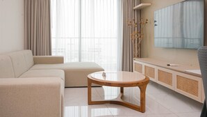 Een moderne woonkamer met een beige bank, een houten salontafel, een televisie op een witte kast en een groot raam met gordijnen.