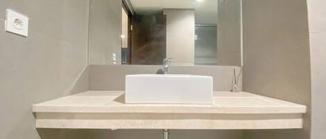 Un baño moderno con un lavamanos rectangular, un espejo y una ducha visible al fondo.