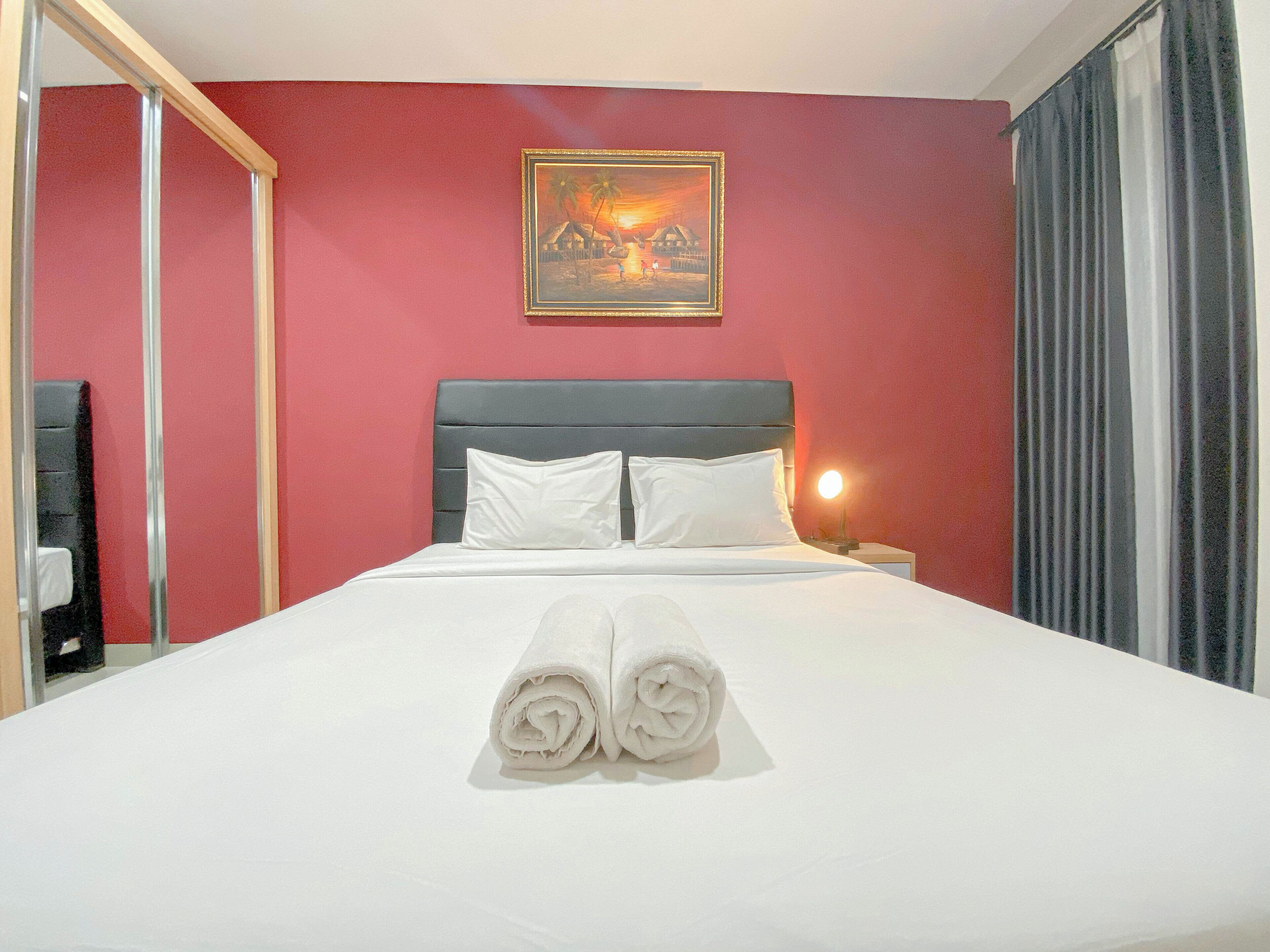 A neatly made bed with white linens and rolled towels, a headboard, a painting of a sunset scene, and a nightstand with a lamp.