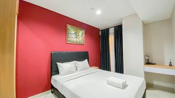 A hotel room with a bed, a red wall, a painting, a window with curtains, and a small table with a lamp.