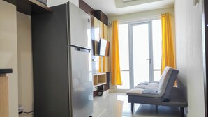 A compact living space with a refrigerator, a flat-screen TV, a sofa, and a sliding glass door with yellow curtains. - Luxurious 1BR at Vida View Makassar Apartment (Makassar)