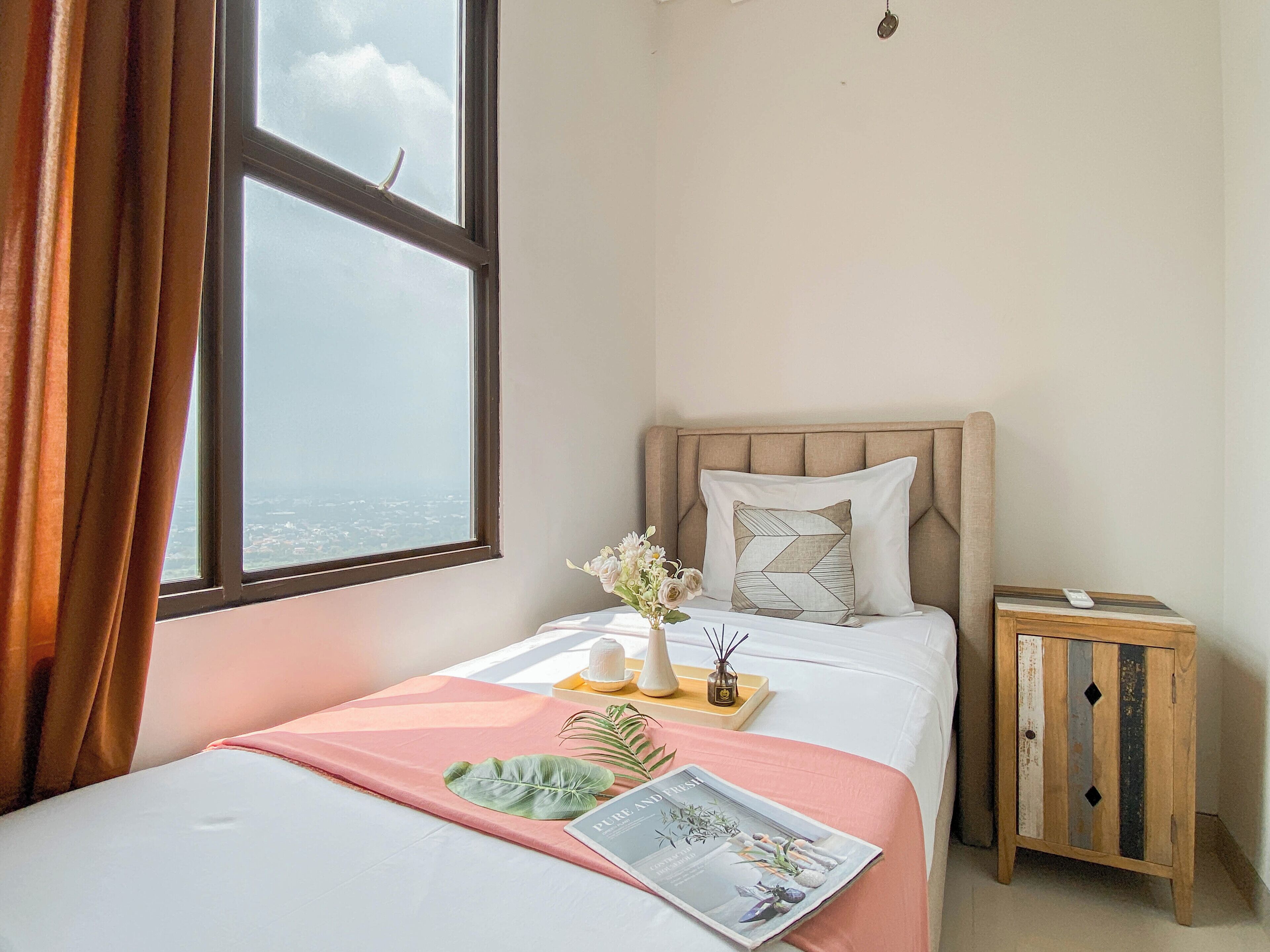 Un lit bien fait, recouvert d’une couvre-lit rose, avec un magazine et une feuille décorative. On aperçoit une table de chevet sur laquelle se trouve un vase de fleurs et une petite lampe. Une fenêtre avec des rideaux est visible, et un meuble en bois est adossé au mur.
