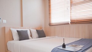 Un lit bien fait, recouvert d’une couverture blanche et bleue, une table de chevet avec une lampe, et une fenêtre avec des stores.