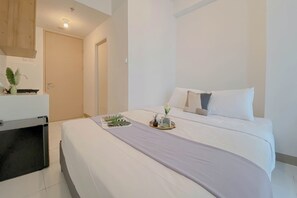 A neatly made bed with a white comforter and a lavender throw, a bedside table with a vase of flowers, and a door leading to another room. - Comfortable Studio Room Apartment Tokyo Riverside PIK 2 (Teluknaga)