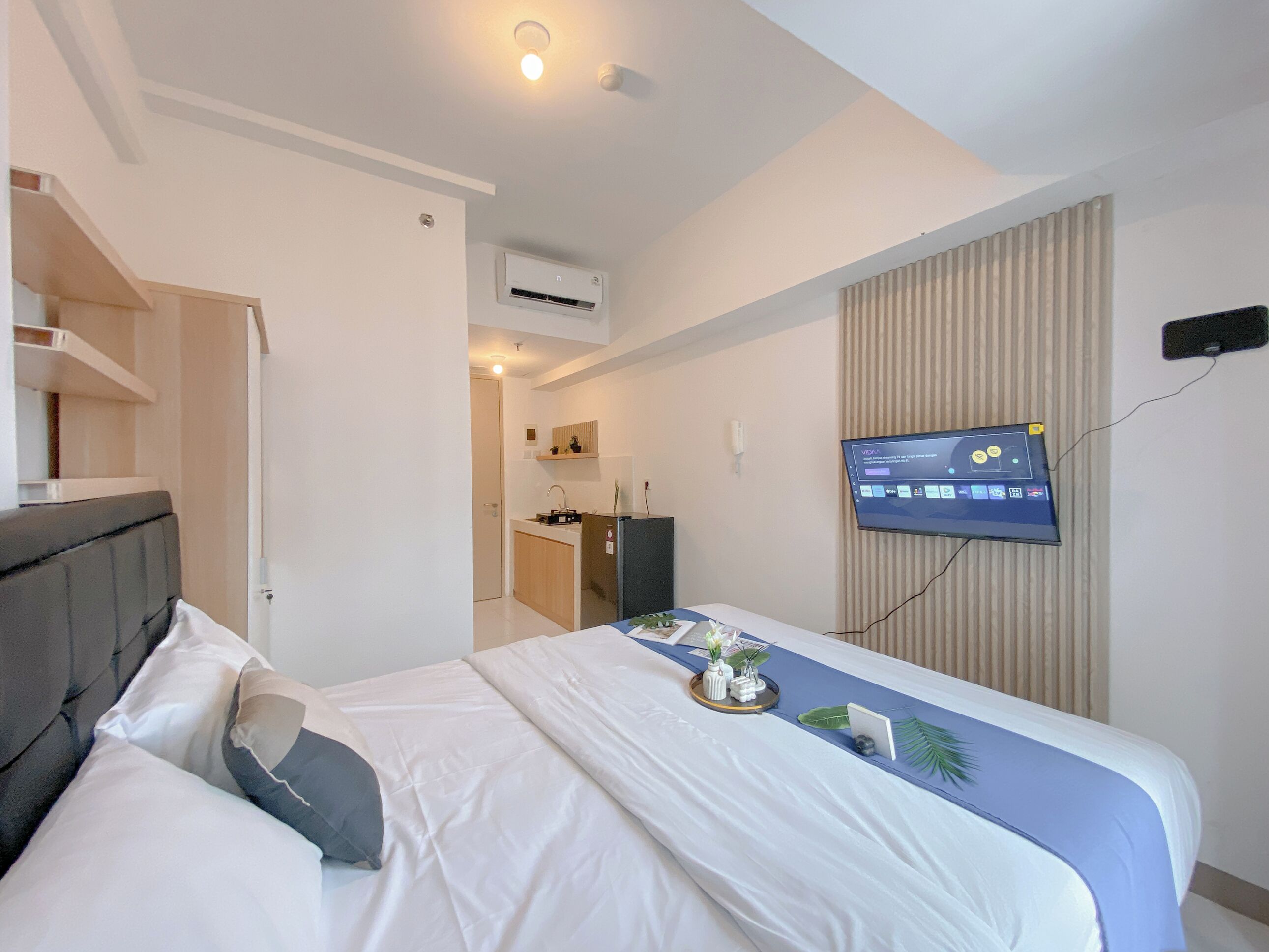 A hotel room with a bed, a TV mounted on the wall, a small kitchenette, and a door leading to another room.