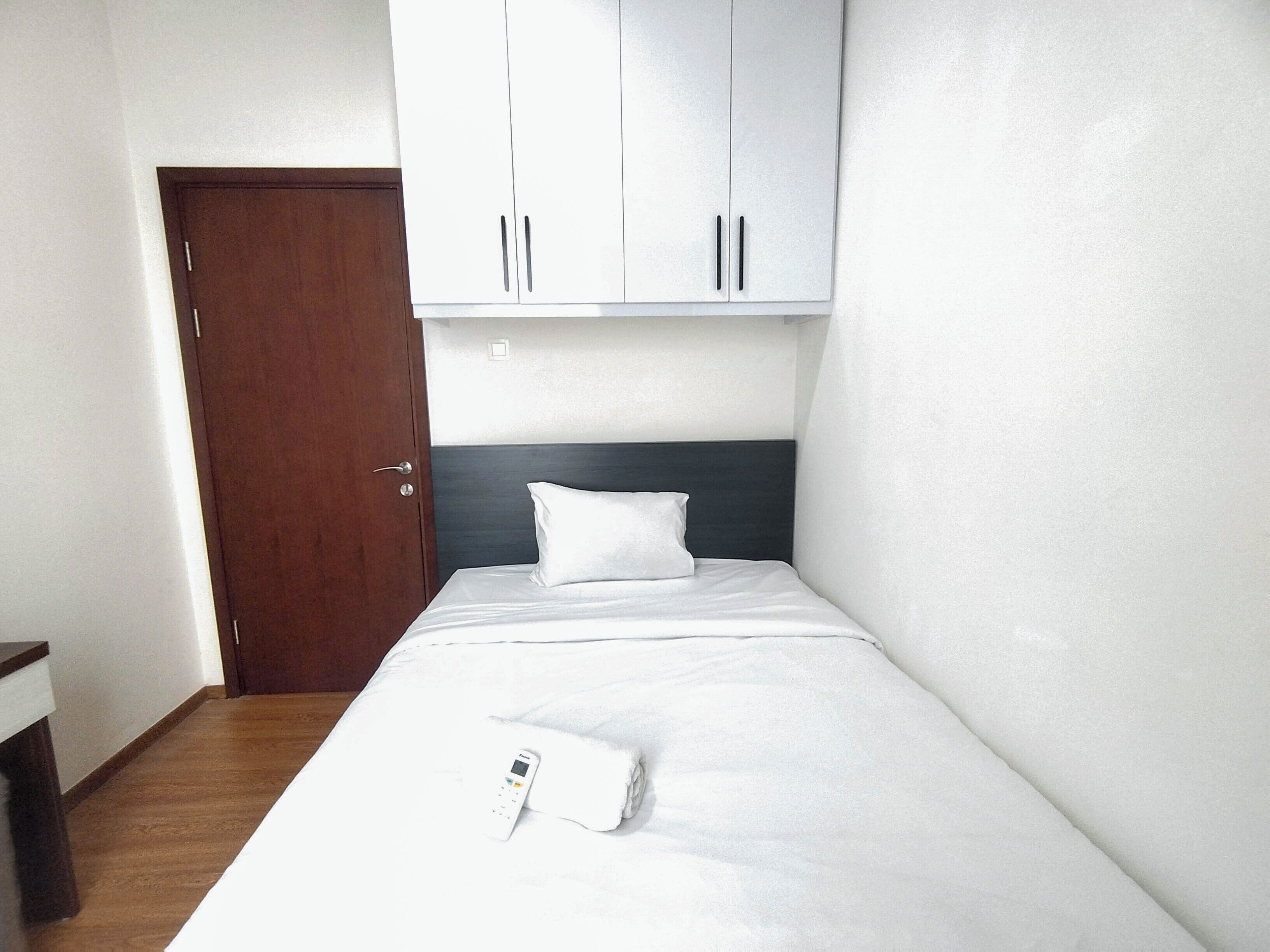A small, neatly made room with a single bed, a wooden door, and white cabinets above the bed.