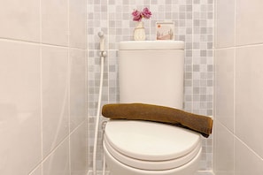 A toilet with a wooden seat cover, a toilet brush, and a small vase with flowers on the tank. - Comfortable and Compact Studio Apartment Tokyo Riverside PIK 2 (Teluknaga)