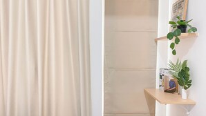Ein Zimmer mit einem beigefarbenen Vorhang, einem hölzernen Hocker, einem an der Wand befestigten Regal mit Pflanzen und einem gerahmten Bild.