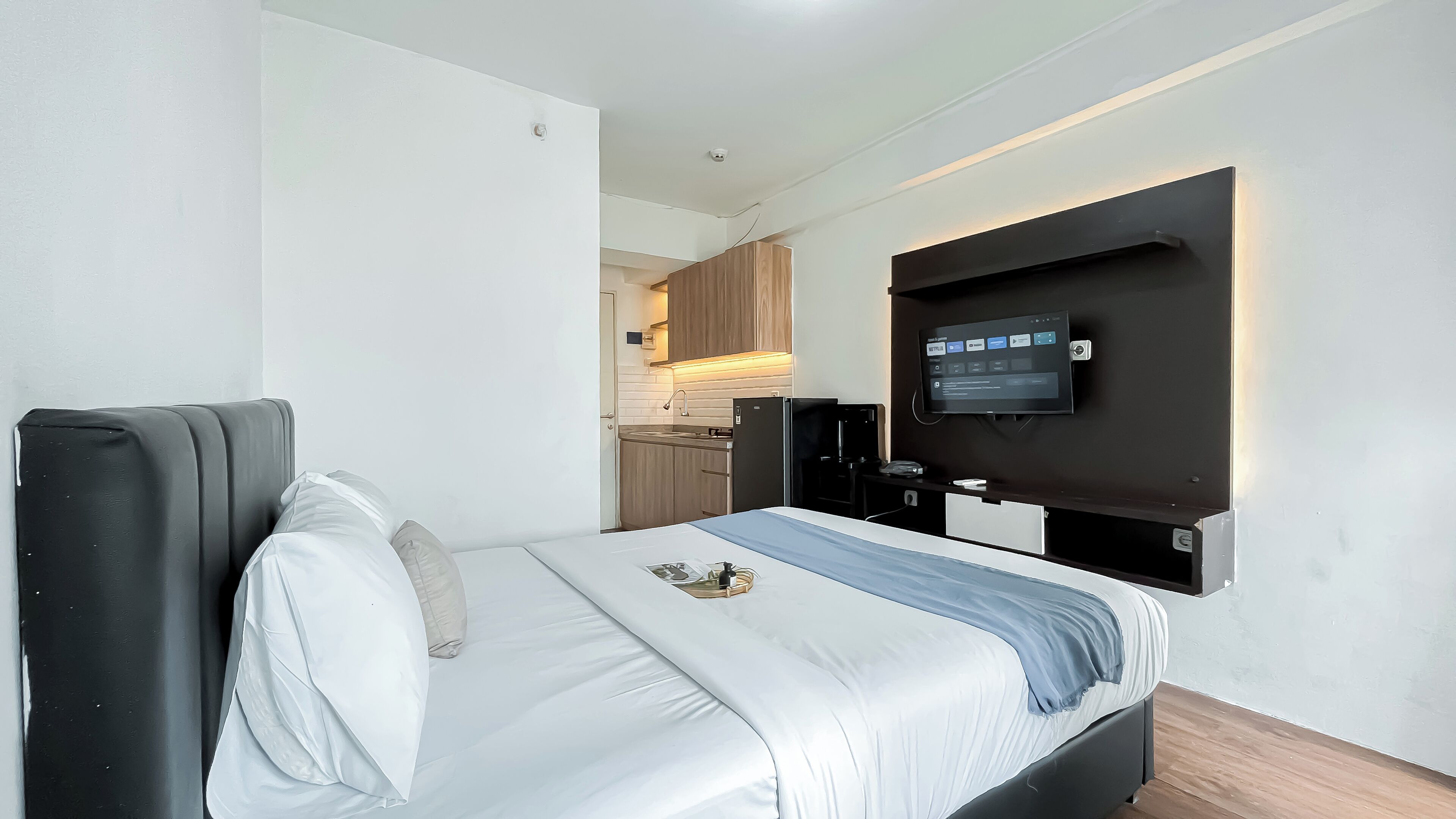 A modern hotel room with a bed, a TV mounted on the wall, and a kitchenette in the background.