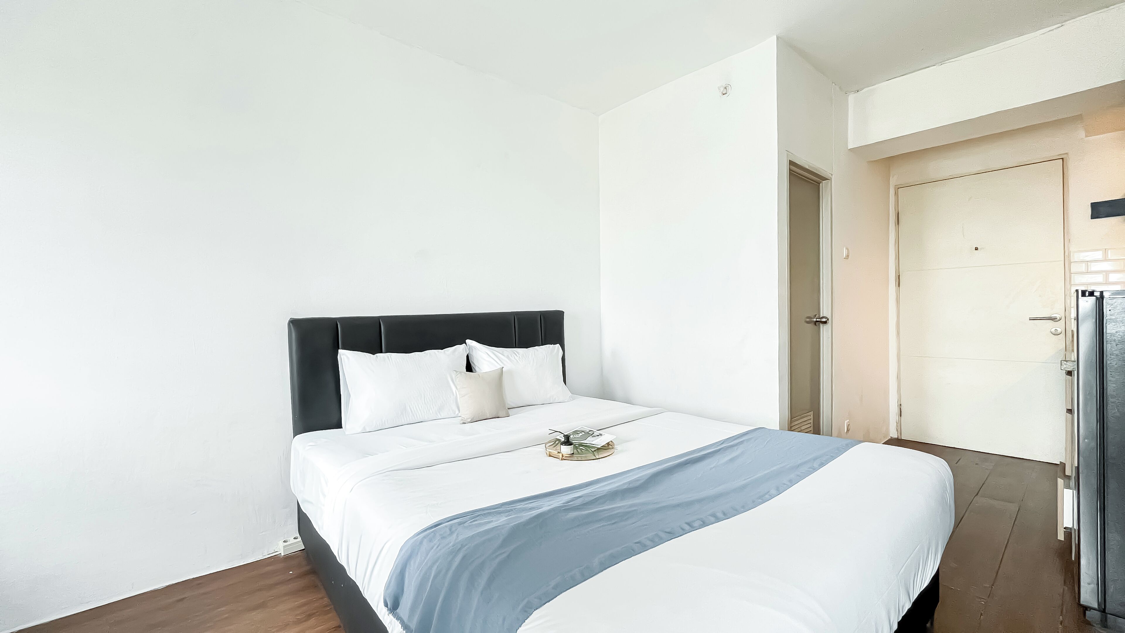 A neatly made bed with a dark headboard and white bedding, a small tray with glasses and a book on the bed, and a door to the right.