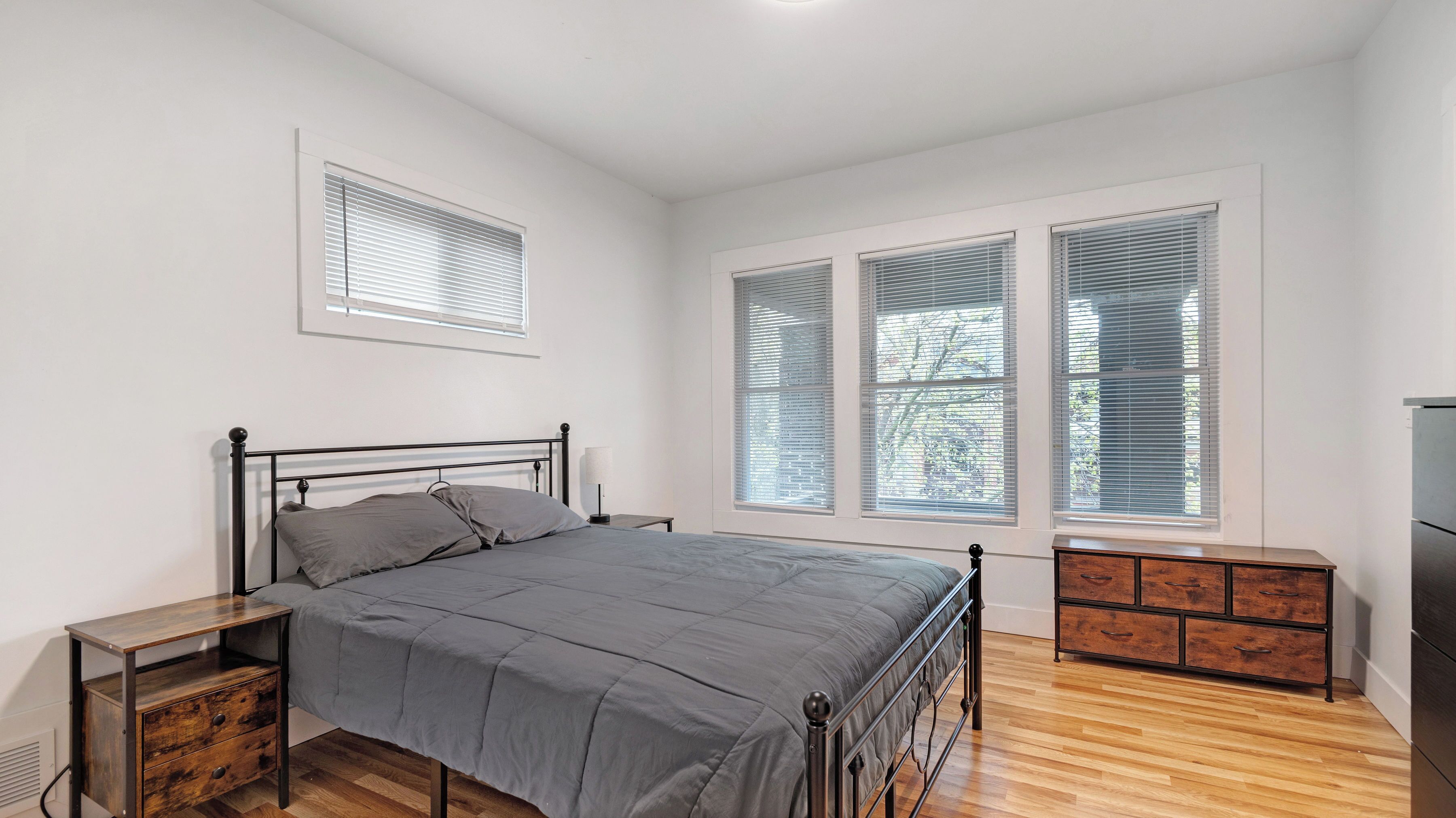 Une chambre à coucher comprenant un lit, des tables de chevet, une commode et de grandes fenêtres équipées de stores.