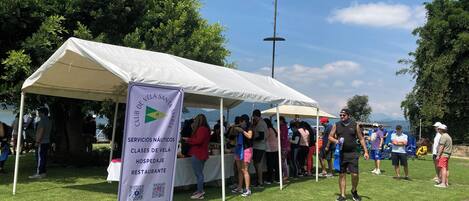 A tent with a banner advertising nautical services and classes, surrounded by people on a grassy area with trees and mountains in the background.