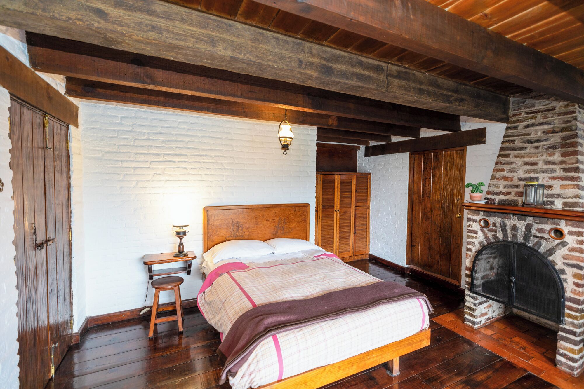 A bedroom with a wooden bed, a nightstand, a stone fireplace, and a white brick wall.