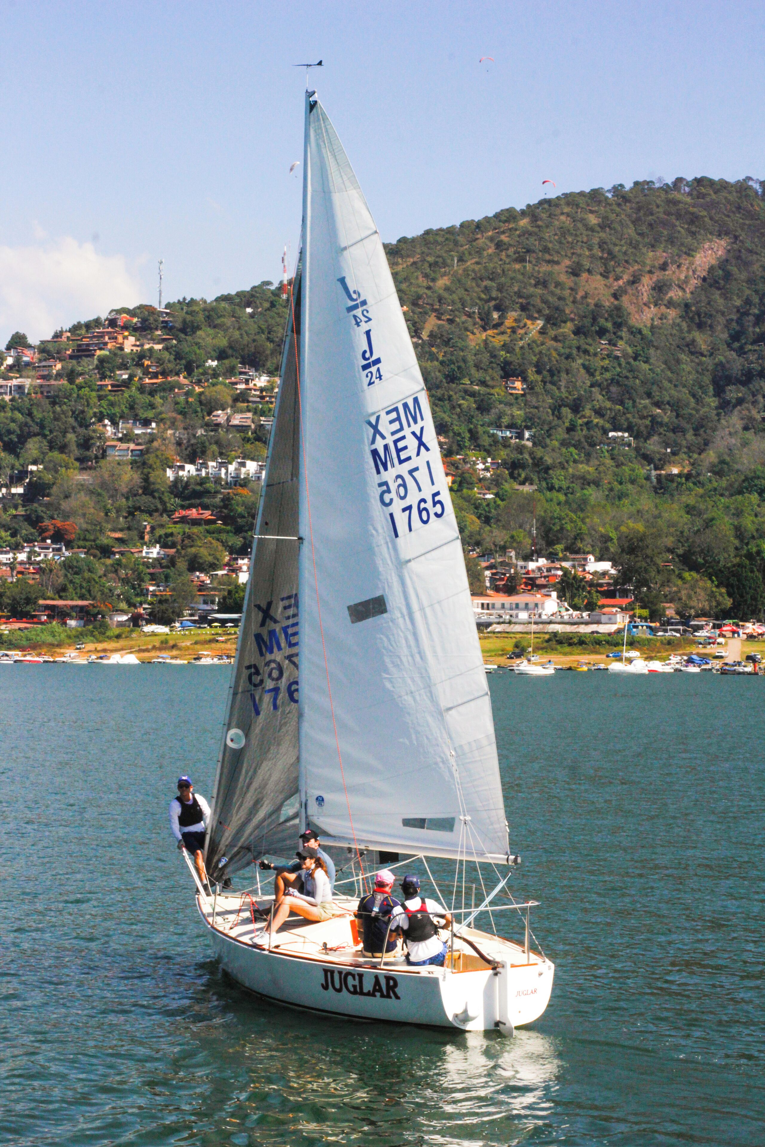 Una barca a vela con lo scritto "JUGLAR" sullo scafo, che naviga in un specchio d'acqua con un paesaggio collinare sullo sfondo.