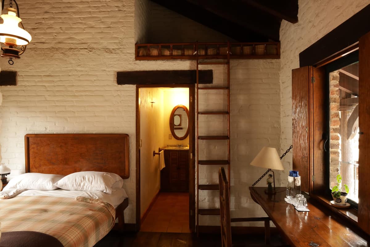 Un dormitorio con una cama de madera, una escalera, un escritorio con una lámpara y una ventana con vistas.