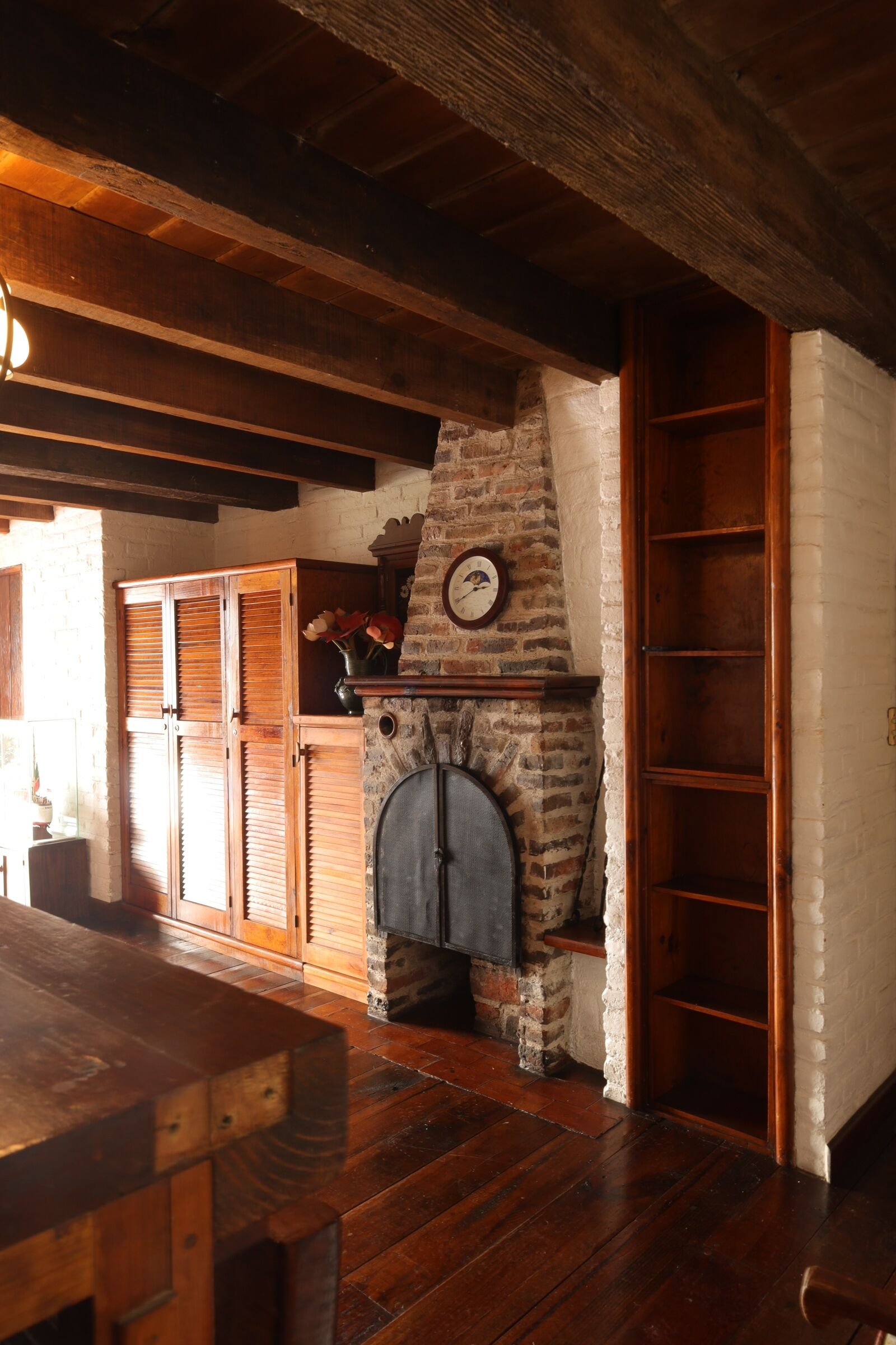 A rustic interior with a wooden table, a brick fireplace, and a wooden shelf with flowers.